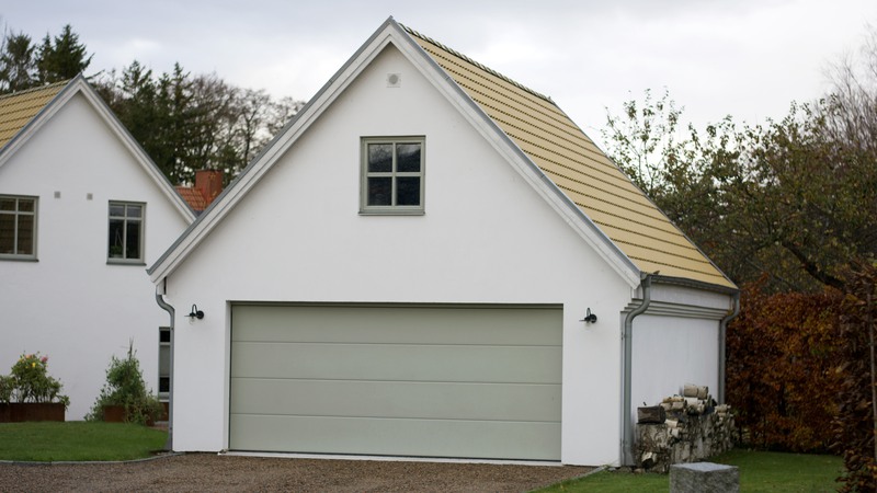 Garagebyggnad med vitputsad fasad, gult tak och ljusgrön garageport. Placerat i anslutning till ett bostadshus.