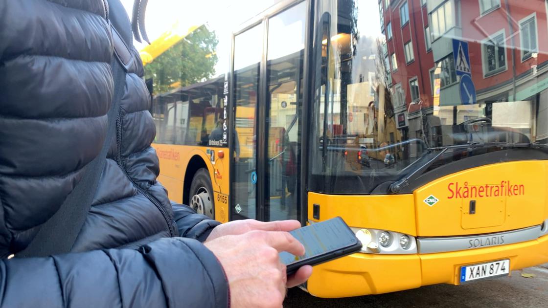 Resenär som kollar busstider i Skånetrafikens app, med gul regionbuss i bakgrunden
