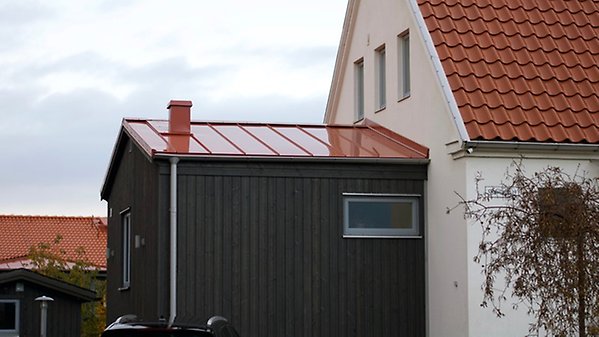 En mindre tillbyggnad med svart träfasad och rött, falsat plåttak på bostadshus med ljus putsfasad och pannbelagt tak.