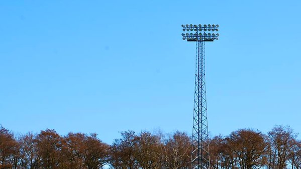 Ljusanordning på mast vid en idrottsanläggning med träd i bakgrunden.